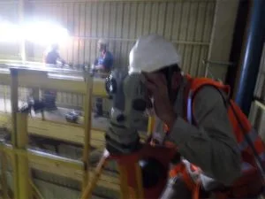 Técnico com capacete branco e colete laranja usa teodolito em inspeção industrial